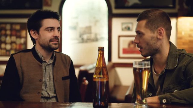 Two old friends are having conversation in pub over beer. Glass and bottle in foreground, tables and pictures on walls in background.