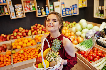 Girl in red holding different fruit and vegetables at basket on fruits store.