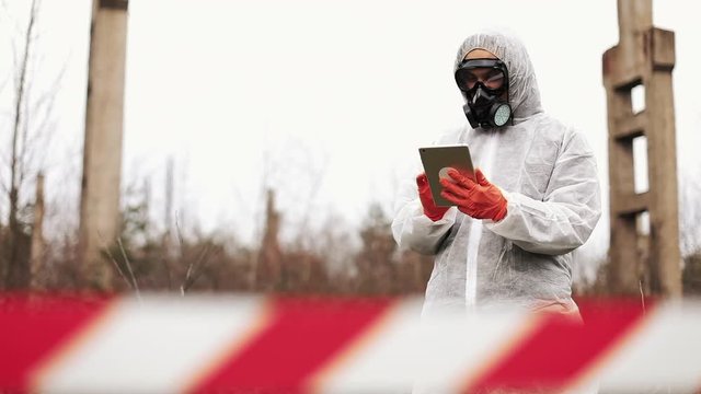 Man in bio-hazard suit and gas mask takes notes in his tablet standing on the polluted land
