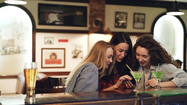 Girls are using smartphone, laughing and talking while sitting in bar together. Girls in casual clothing are watching screen and chatting. Modern technologies for fun concept.