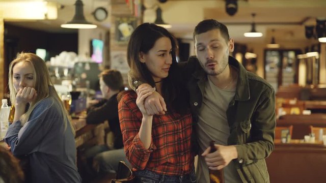 Dolly shot of young woman helping her drunk boyfriend leave bar. Handsome bearded man is holding beer bottle and telling something to his girlfriend.