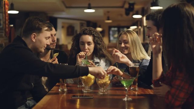 Group of friends are drinking, eating snacks and talking while sitting at table in bar. Young people enjoying themselves and communicating in pub concept.