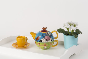 Tetera decorada, taza de té y maceta en  bandeja blanca de madera.