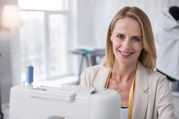 Customer oriented focus. Positive female tailor grinning to camera while using sewing machine