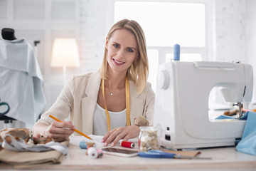 Exceptional talent. Exuberant female couturier smiling to camera while sketching