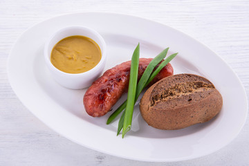 Beef sausage with mustard sauce on a white plate, green onions and black bread on a white wooden background
