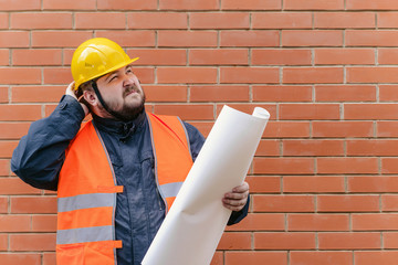 Engineer in a helmet at the construction site against a brick wall is with drawings and surprised scratching his head.
