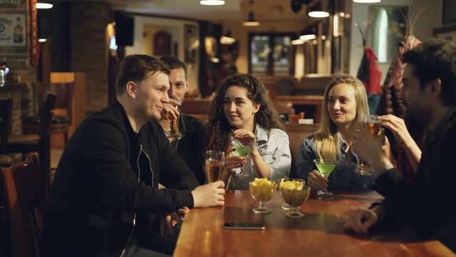 Young people are celebrating something in cafe. They are clinking glasses with beer and cocktails, drinking and chatting. Modern celebration in bar concept.