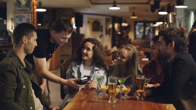 Cheerful students are paying with smartphone while sitting at table in nice cafe. Quick easy contactless payment concept. Attractive people and modern bar interior are visible.