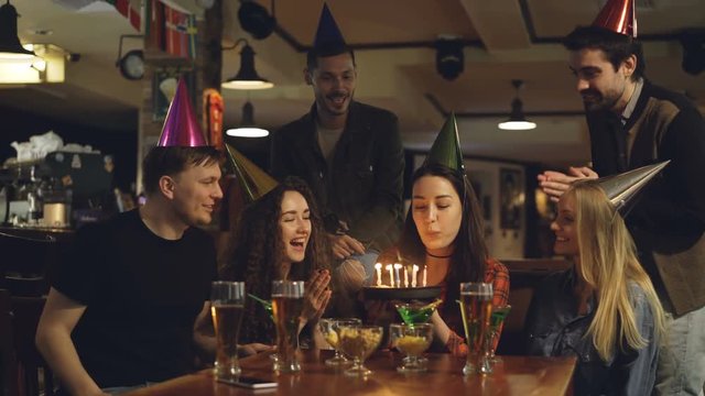 Young pretty brunette is making wish and blowing out candles on birthday cake while celebrating birthday in cafe with friends. Happy people in party hats are clapping hands.