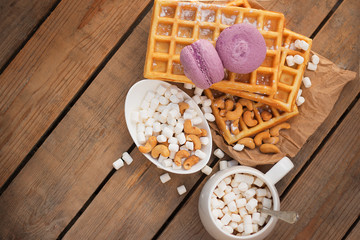 Belgian waffles, a cup of coffee and marshmallows, macaroons, cashew nuts on a wooden table. Top view