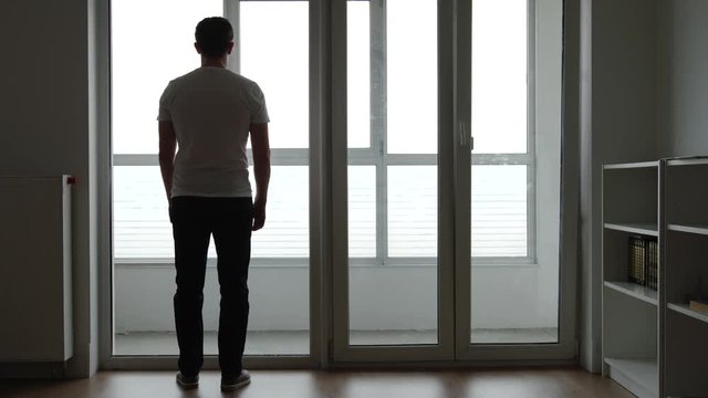 Silhouette of sad young adult stands near a big window at home