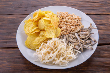 Dried squids, potato chips, the dried small fish, nuts peanut on a white plate and a dark wooden background