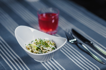 Table in the restaurant. On the table is a salad in a white plate, a knife, a fork, a glass with red juice. On the table is a blue tablecloth in strips.