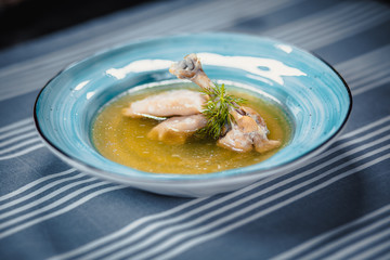 Soup in blue plate with chicken broth, chicken's leg, dumplings and greenery. Plate on the table with a blue tablecloth in white strips.