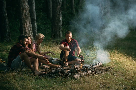 Two couples camping in woods. Cooking sausages on campfire. Warm summer evening in forest