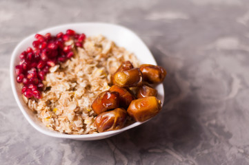 Cooked yummy warm oatmeal and heap of ripe fresh pomegranate seeds and lot of dried dates in white ceramic bowl on worn gray scratched concrete with copy space