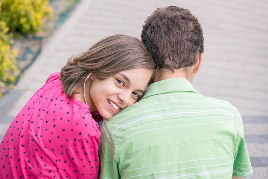 Happy teenage boy and girl with headphones are using gadget, talking and smiling while sitting on the stairs outdoors. Young sister and brother teens playing on mobile phone and listening to music.