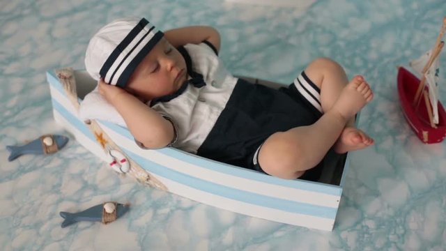 Cute toddler baby boy sleeping in wooden boat with fishes, starfish and binoculars, dressed as sailman, indoors shot