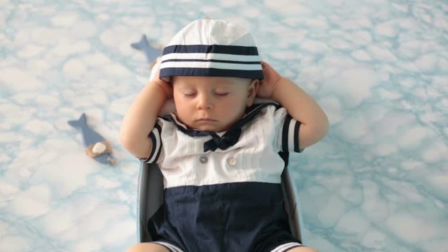 Cute toddler baby boy sleeping in wooden boat with fishes, starfish and binoculars, dressed as sailman, indoors shot