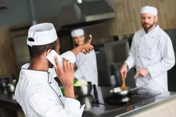 Fototapeta na wymiar african american chef talking by smartphone and showing thumb up to colleagues at restaurant kitchen