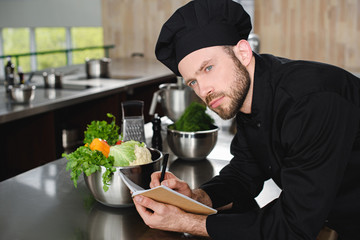 handsome chef writing down new recipe to notebook at restaurant kitchen and looking away