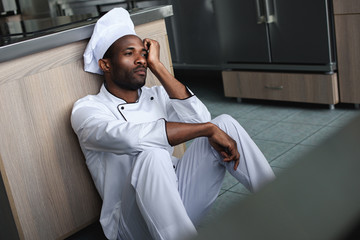 tired african american chef sitting on floor at restaurant kitchen and looking away
