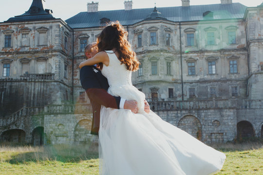 Wedding couple is walking outdoors in sunny day. Medieval castle stone walls with old renaissance palace on background. Groom is hugging bride is satin lace dress. Old architecture and green grass