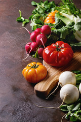 Variety of wet raw fresh organic colorful vegetables tomatoes, radish with leaves, fennel, paprika, salt for salad on wooden chopping board over dark brown texture background. Close up, copy space.