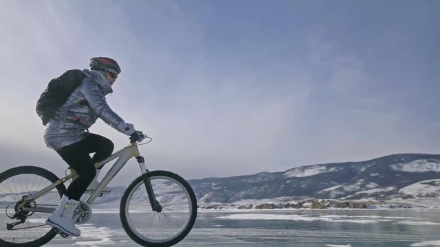 Woman is riding bicycle on the ice. The girl is dressed in a silvery down jacket, cycling backpack and helmet. Ice of the frozen Lake Baikal. The tires on the bicycle are covered with special spikes