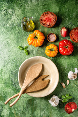 Variety of red and yellow organic tomatoes with olive oil, garlic, salt, empty ceramic bowl, wooden spoons for salad over green texture background. Top view, space.