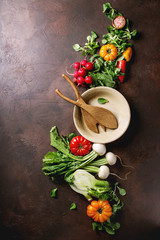 Variety of wet raw fresh organic colorful vegetables tomatoes, radish with leaves, fennel, paprika, salt, ceramic bowl and wood spoons for salad over dark brown texture background. Top view, space.