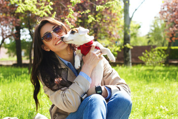 Naklejka premium Portrait of attractive young woman hugging cute jack russell terrier puppy in park, green lawn, foliage background. Hipster female in sunglasses smiling, pets her dog on grass. Close up, copy space.