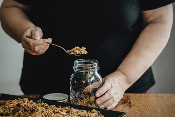 Woman filling a mason jar with homemade granola. Healthy vegan snack easily prepared at home. Visible body parts, hands of an elderly woman.
