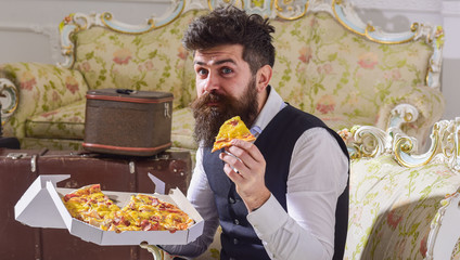 Macho in classic clothes hungry, holds slice of cheese pizza, eats, enjoying taste, interior background. Man with beard and mustache holds box with tasty fresh hot pizza. Bon appetit concept.