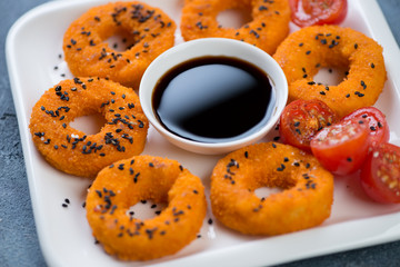 Closeup of roasted calamari rings with black sesame seeds and soy sauce on a white plate, selective focus