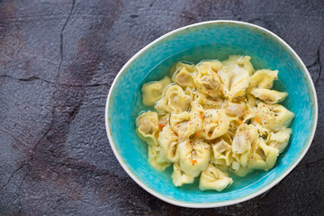 Blue bowl with tortellini in bouillon, high angle view on a weathered asphalt background, horizontal shot with copyspace