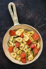 Tortellini with fried zucchini and cherry tomatoes in a serving pan, flat-lay on a dark brown stone background, vertical shot