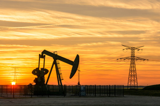 Pumpjack And Transmission Towers At Sunset Symbolizing Energy Transition. A Pump Jack Pumping Oil Out Of A Well With Silhouettes Of Electricity Pylons And Power Line Against A Red Sky.