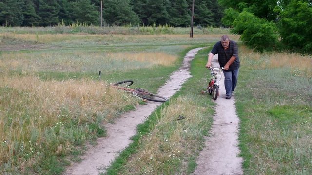 Fat senior man walks on a country road with kids tricycle and found an old broken bicycle he likes