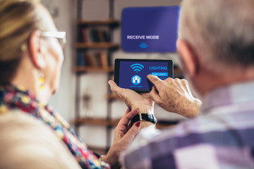 Elderly couple sitting comfortably on a sofa with their backs holding remote home control system on a digital tablet - Powered by Adobe
