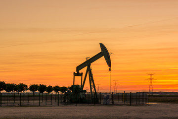 Pumpjack and transmission towers at sunset symbolizing energy transition. A pump jack pumping oil out of a well with a row of trees and a row of electricity pylons against a red sky.