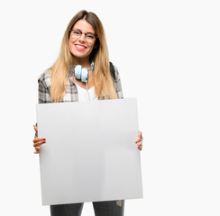 Fototapeta premium Young student woman with headphones and backpack holding blank advertising banner, good poster for ad, offer or announcement, big paper billboard