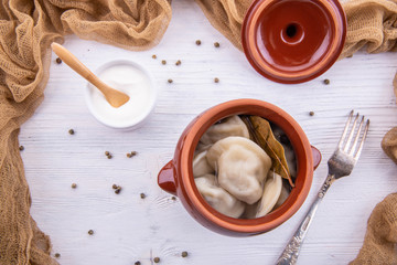 Pelmeni in a pot with spices and sour cream on a white wooden background