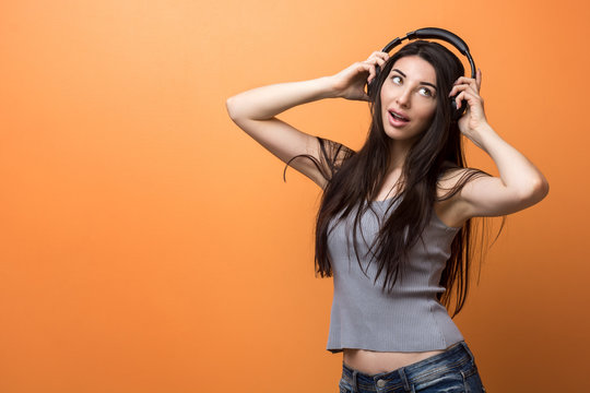 Portrait of a young girl listening to the music through the headphones and holding it in her hands against orange background. Lifestyle, people and technologies concept