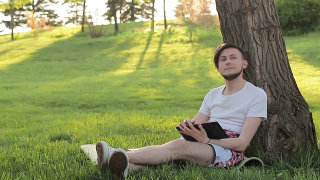 Satisfied man sits under a tree and uses a tablet