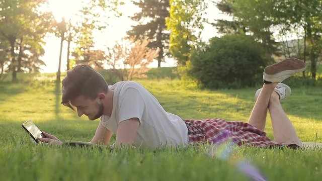 A young man lies on the grass with a tablet communicating with someone on the web