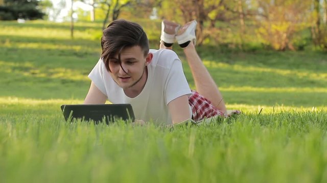 A young man lies on the grass with a tablet communicating with someone on the web
