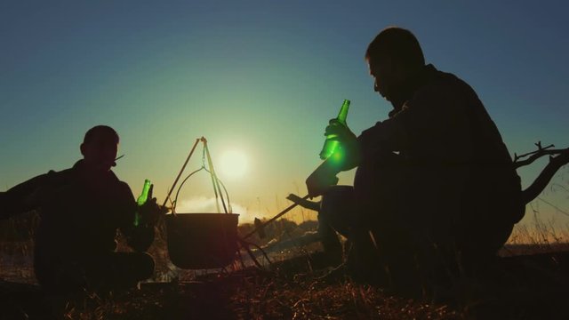 Two fellow campers preparing food in the forest. Two cheerful friends by the bonfire at sunset. dventure, travel, tourism and people concept.