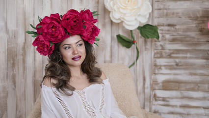 Girl model posing. a young woman in a wreath of scarlet peonies on her head, dark long curly hair descends on small shoulders